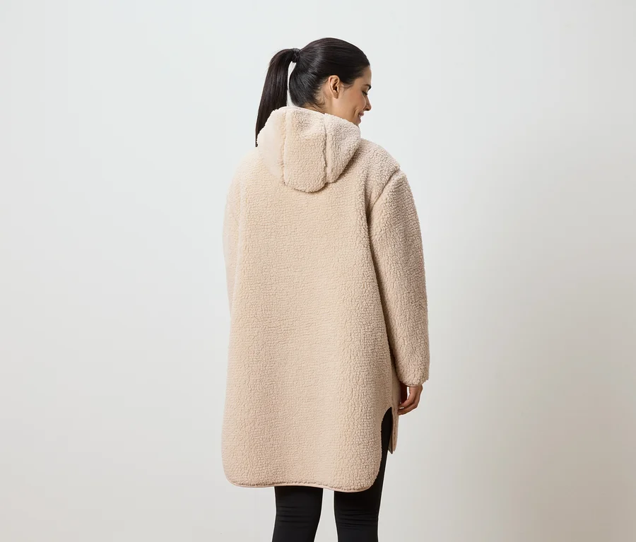 Une femme portant une queue de cheval porte un long manteau en peluche beige à capuche et des leggings noirs, vue de dos.