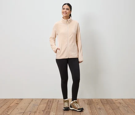 Femme portant une veste en polaire beige zippée, un legging noir et des chaussures de randonnée marron devant un mur blanc.
