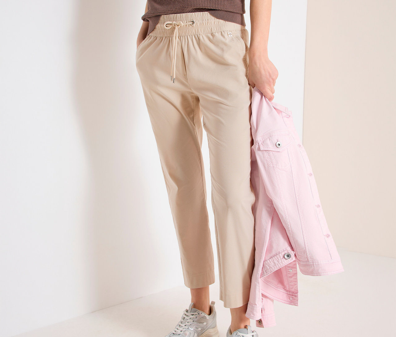Femme portant un pantalon de jogging CECIL « Tracey », un haut marron et des baskets argentées. Elle tient une veste rose.