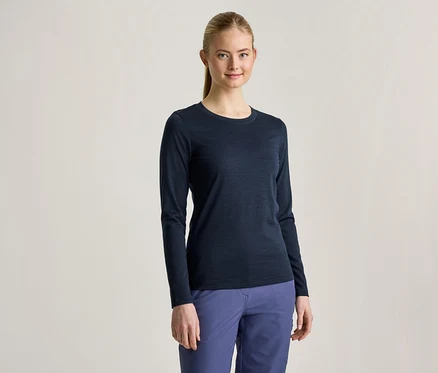 Une jeune femme aux cheveux blonds attachés en queue de cheval porte un t-shirt à manches longues bleu foncé et un pantalon bleu, elle sourit à la caméra.