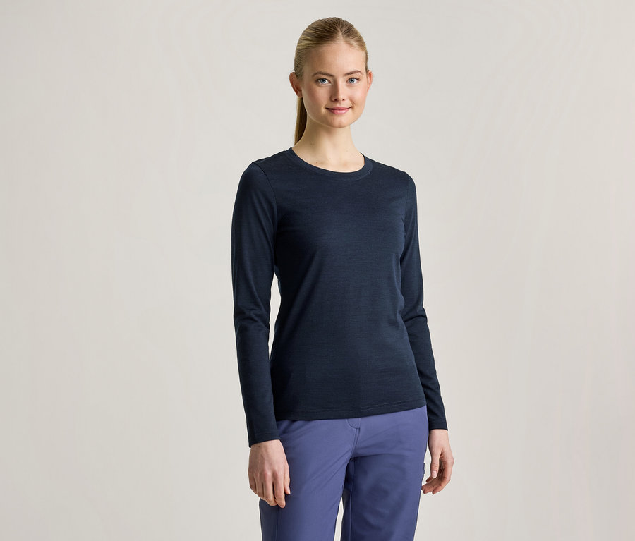 Une jeune femme aux cheveux blonds attachés en queue de cheval porte un t-shirt à manches longues bleu foncé et un pantalon bleu, elle sourit à la caméra.