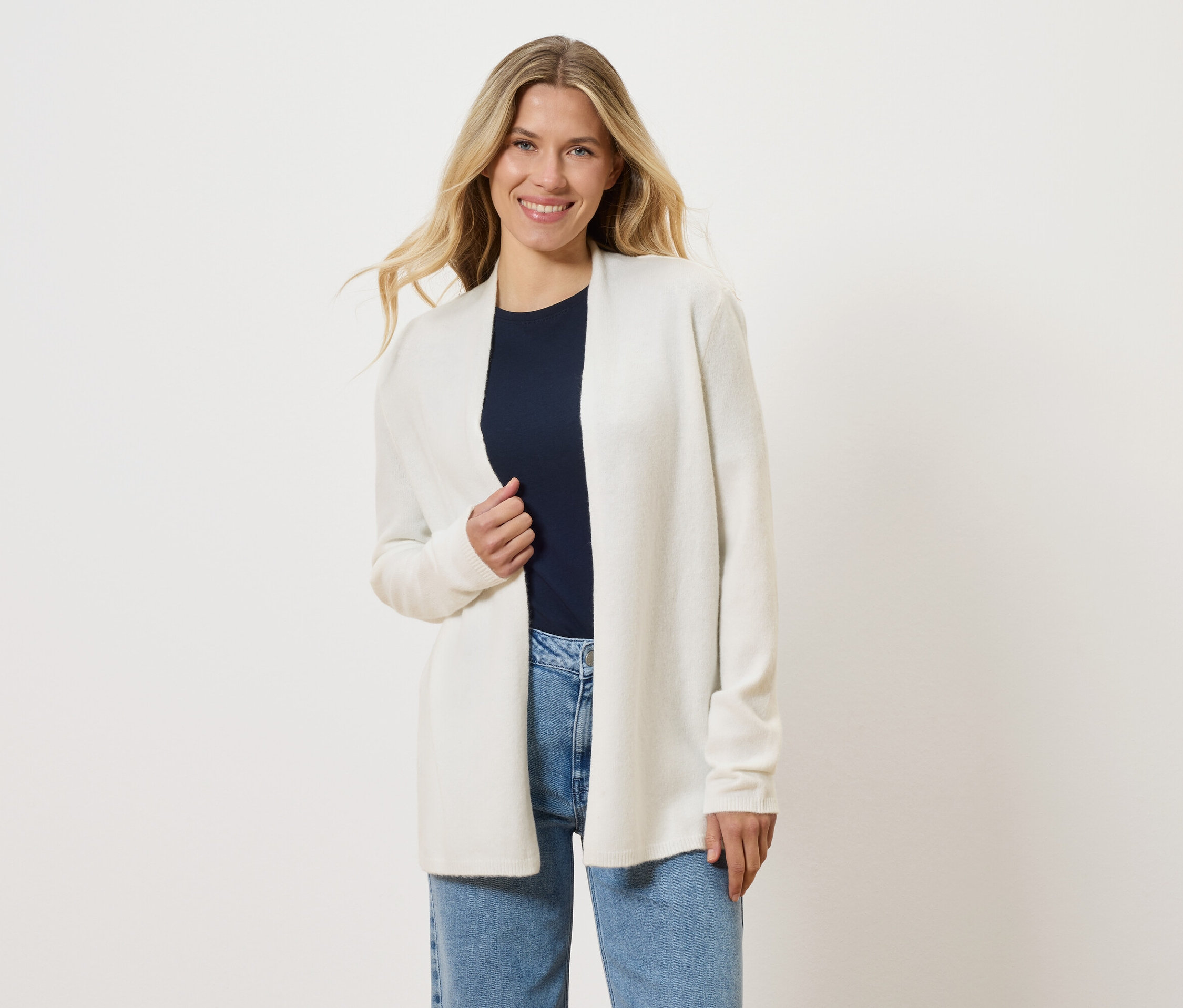 Une femme blonde porte un cardigan ouvert blanc sur un t-shirt bleu marine et un jean bleu.
