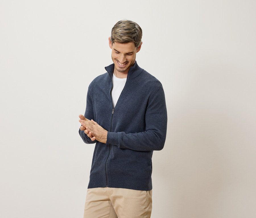 Un homme sourit et se frotte les mains. Il porte un cardigan bleu marine et un pantalon beige.