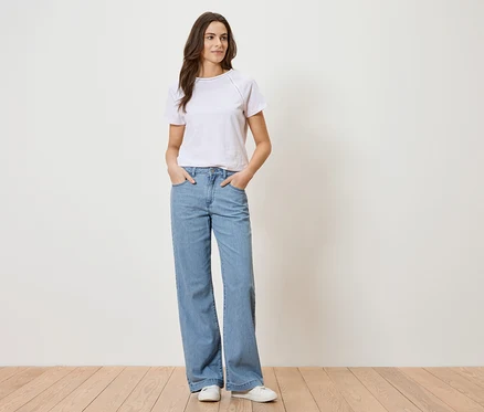 Femme portant un t-shirt blanc et un jean bleu debout devant un mur blanc.