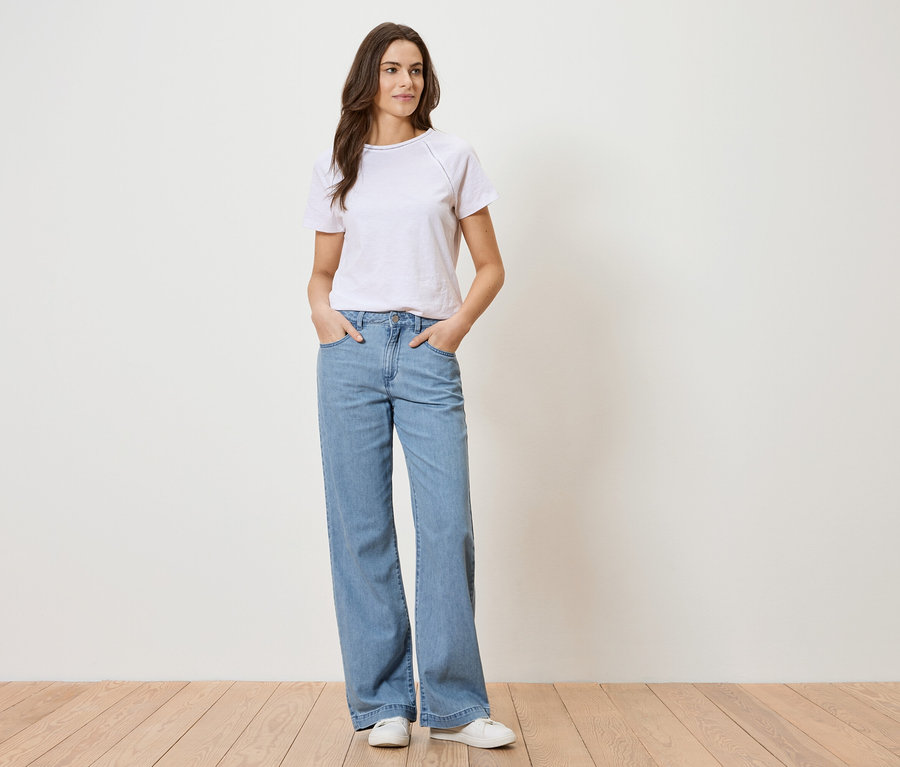 Femme portant un t-shirt blanc et un jean bleu debout devant un mur blanc.