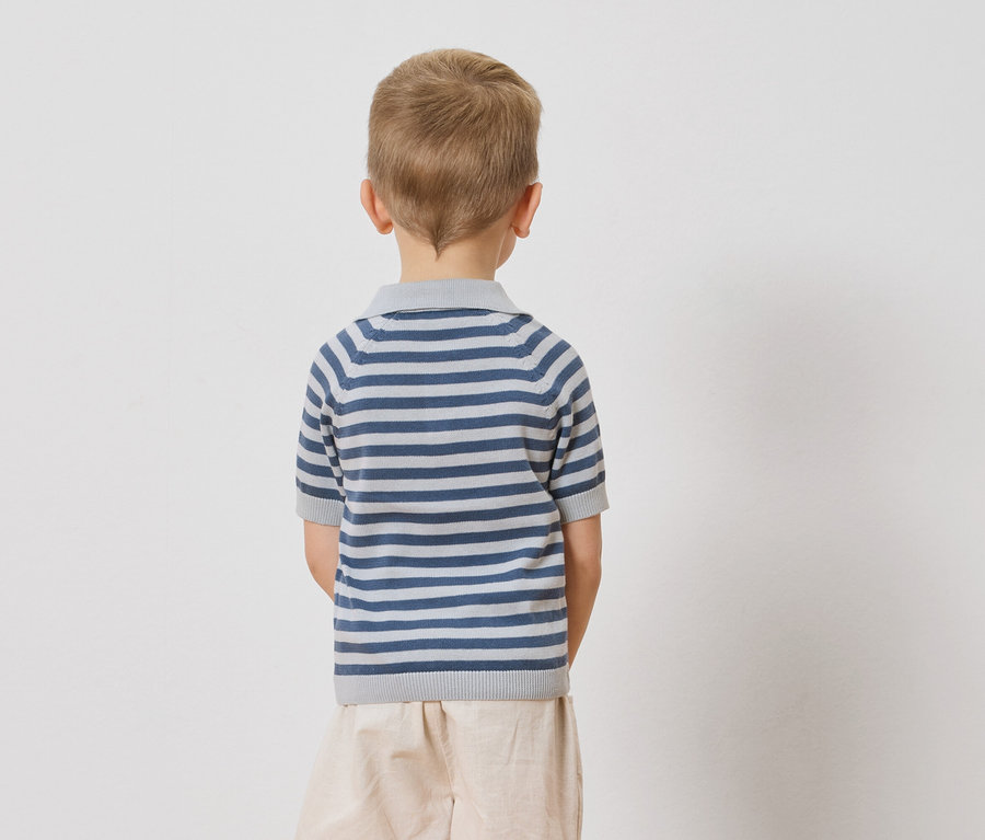 Vue de dos d'un enfant aux cheveux blonds portant un polo rayé bleu et blanc et un short beige sur un fond blanc.