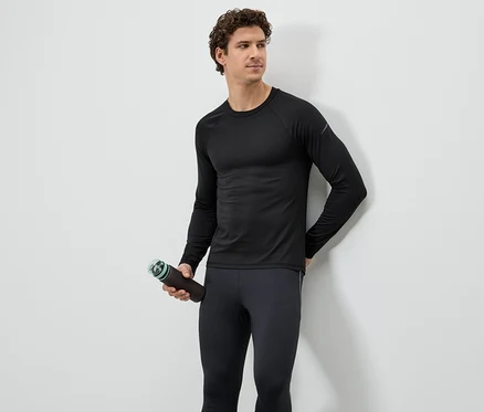 Un homme portant un haut de sport noir et des leggings tient une bouteille d'eau marron devant un mur blanc.