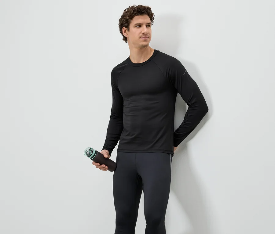 Un homme portant un haut de sport noir et des leggings tient une bouteille d'eau marron devant un mur blanc.