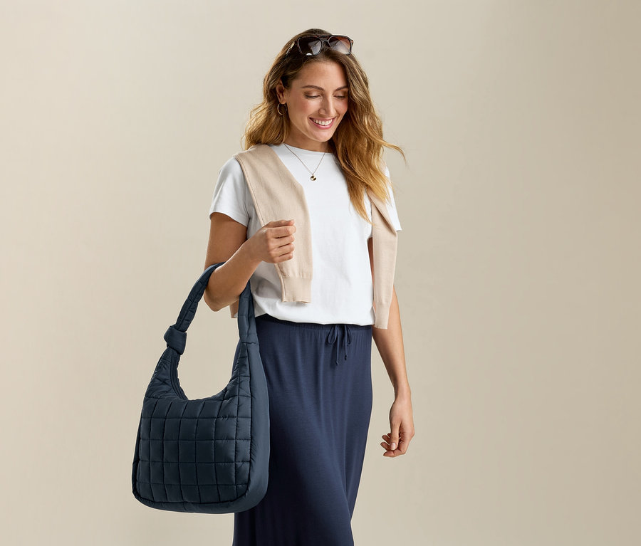 Femme avec des lunettes de soleil sur la tête, un pull beige, un t-shirt et une jupe longue en jersey, tenant un sac bleu.