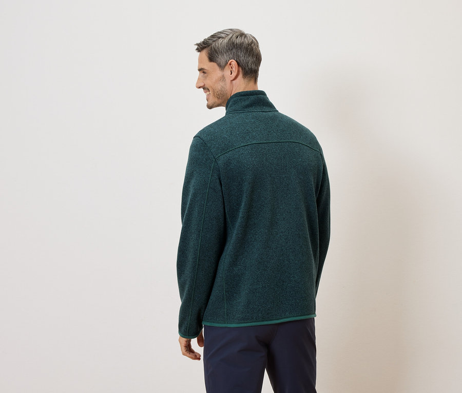 Un homme aux cheveux gris, vu de dos, porte une veste polaire verte et un pantalon bleu marine sur un fond crème.