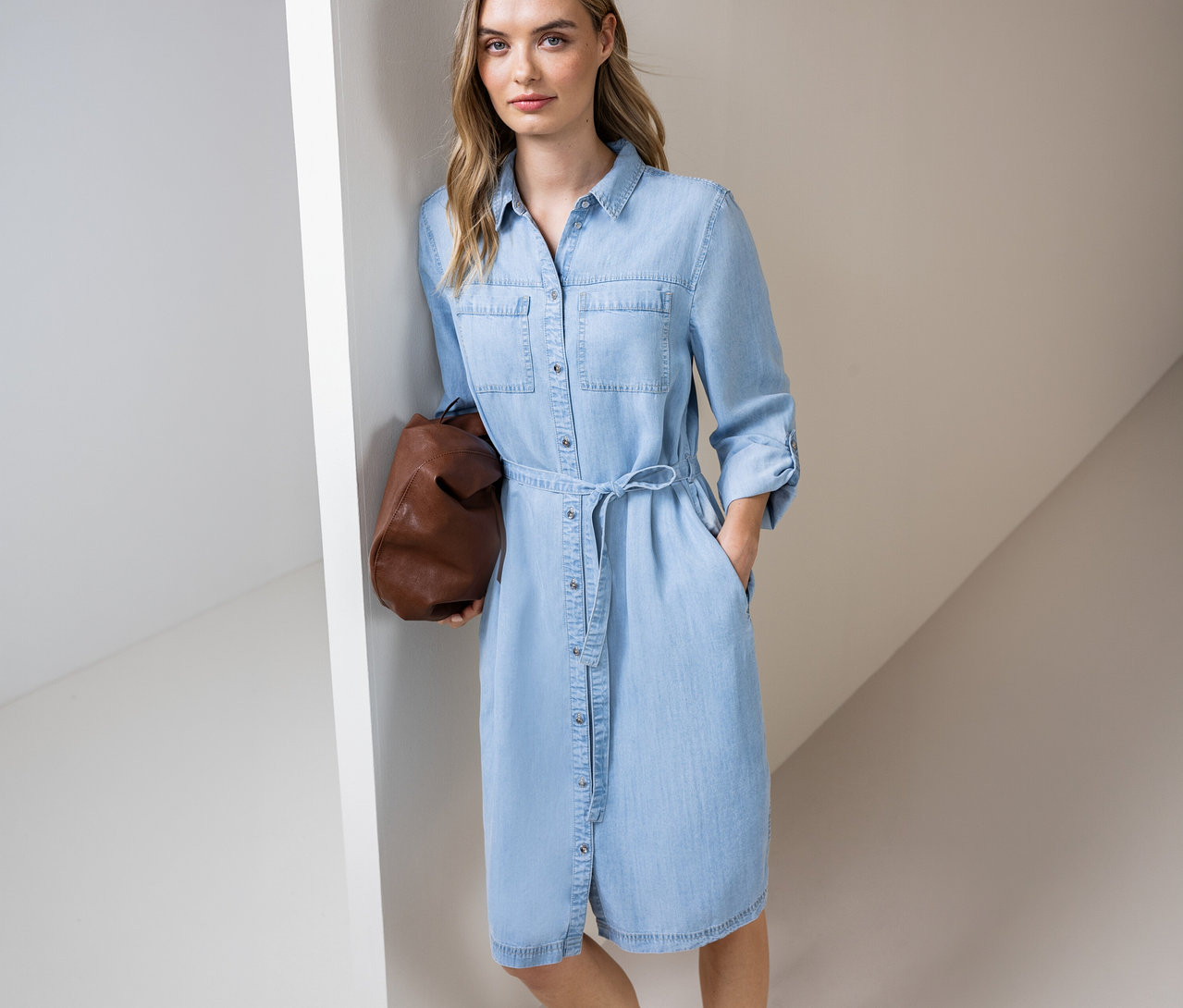 Une femme portant une robe en jean bleu clair avec une ceinture et un sac marron, debout contre un mur.