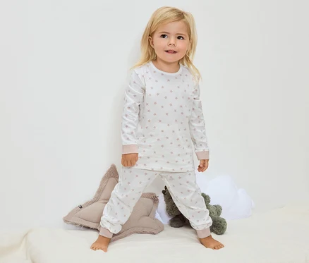 Une petite fille blonde se tient pieds nus sur une couverture, portant un pyjama blanc à motifs étoilés.