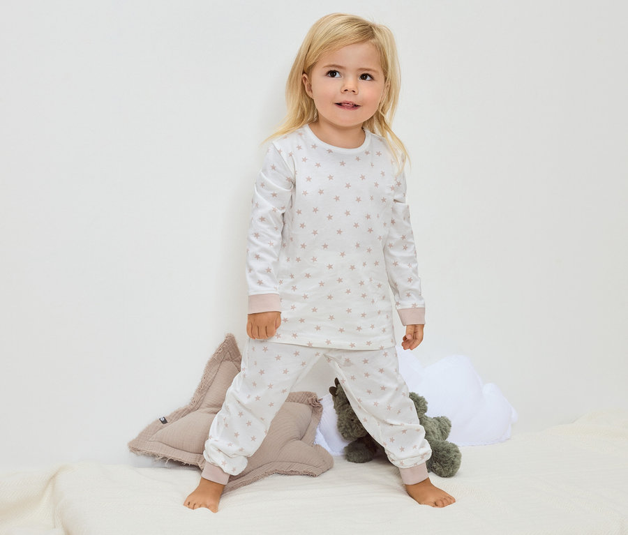 Une petite fille blonde se tient pieds nus sur une couverture, portant un pyjama blanc à motifs étoilés.