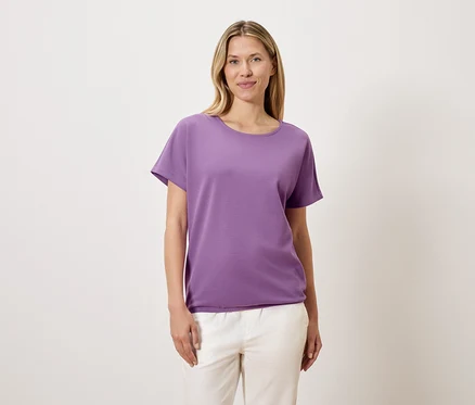 Une femme aux cheveux blonds porte un t-shirt violet et un pantalon blanc.