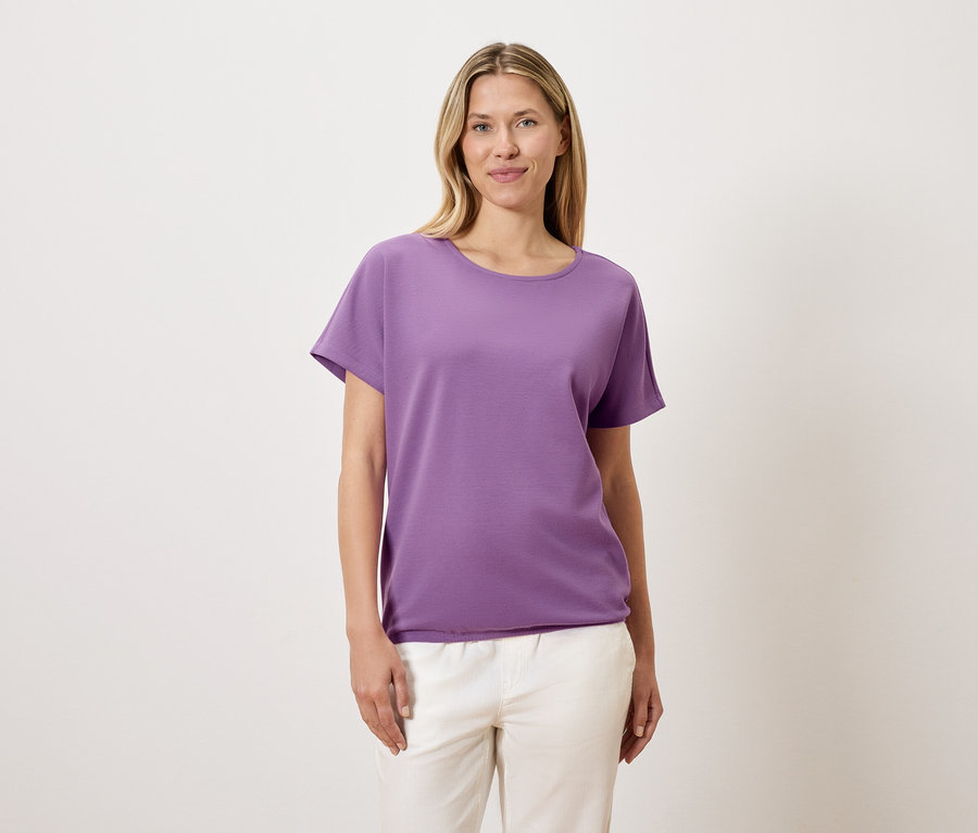 Une femme aux cheveux blonds porte un t-shirt violet et un pantalon blanc.