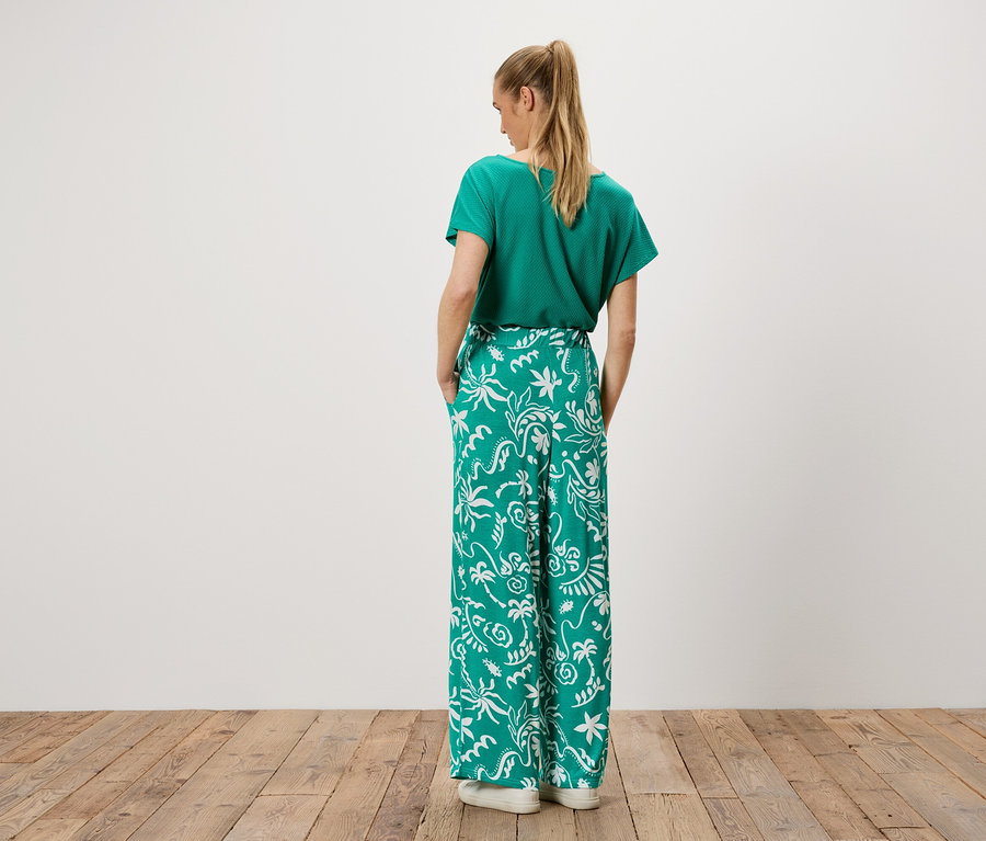 Une femme se tient dos à la caméra, elle porte un t-shirt vert et un pantalon vert avec un motif blanc.