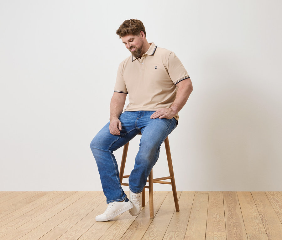 Un homme barbu est assis sur un tabouret, portant un polo beige, un jean bleu et des baskets blanches.