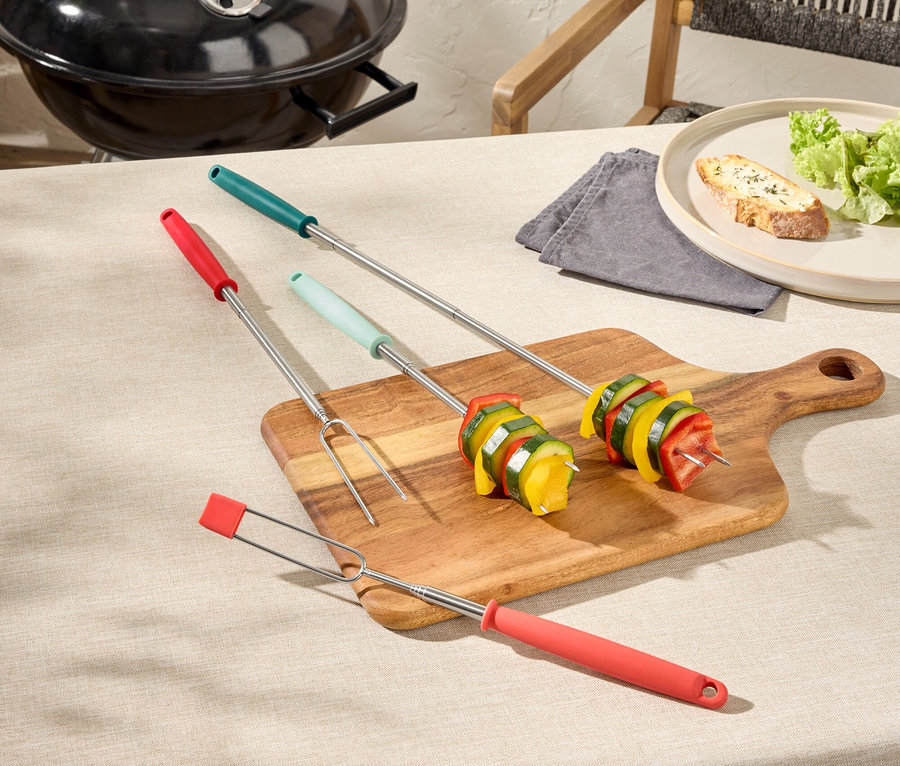 Trois brochettes de barbecue avec des légumes sur une planche à découper en bois, un barbecue et une assiette de pain en arrière-plan.