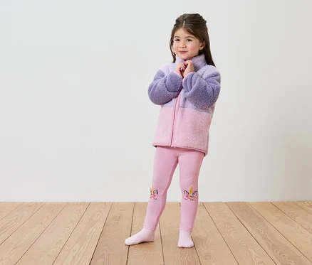 Une fille portant une veste en polaire violette et rose et un legging rose avec un motif de licorne se tient sur un plancher en bois devant un mur blanc.