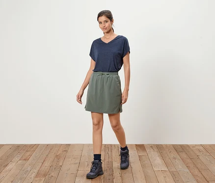 Une femme se tient sur un plancher en bois, portant un t-shirt bleu marine, une jupe vert olive et des bottes bleu marine.