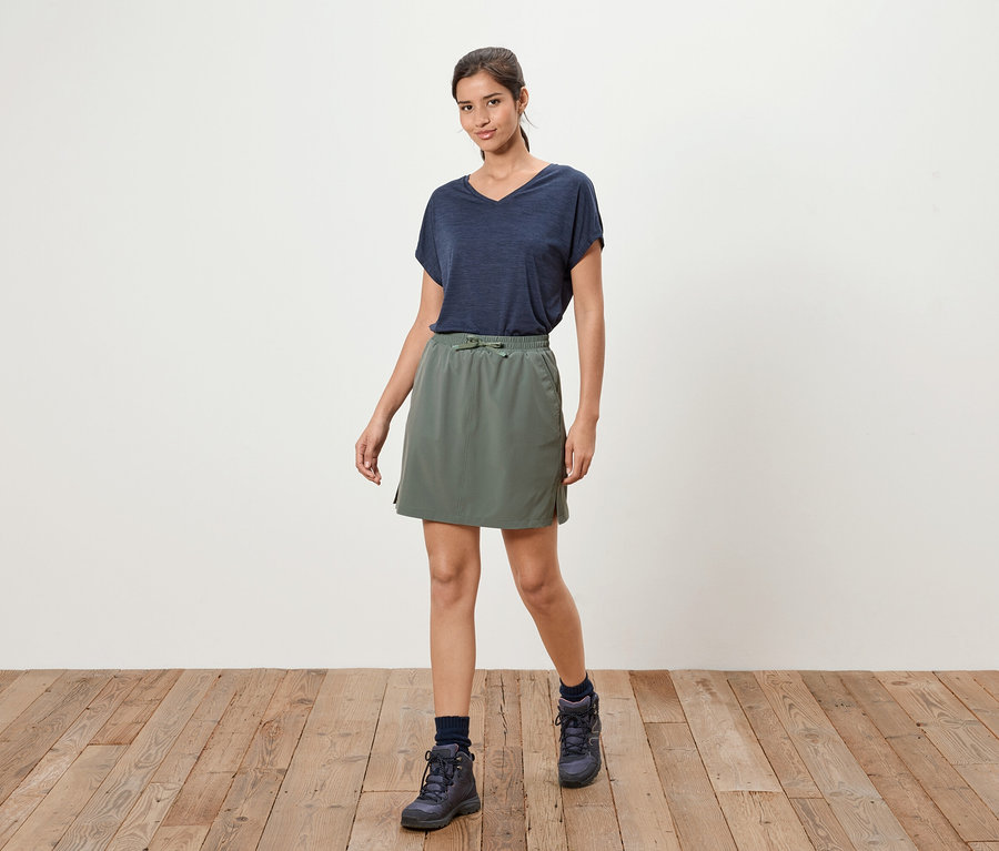 Une femme se tient sur un plancher en bois, portant un t-shirt bleu marine, une jupe vert olive et des bottes bleu marine.