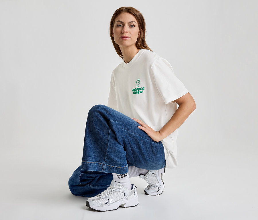 Une femme en jean et t-shirt avec l'inscription "Coffee Crew" est accroupie et porte des baskets blanches.