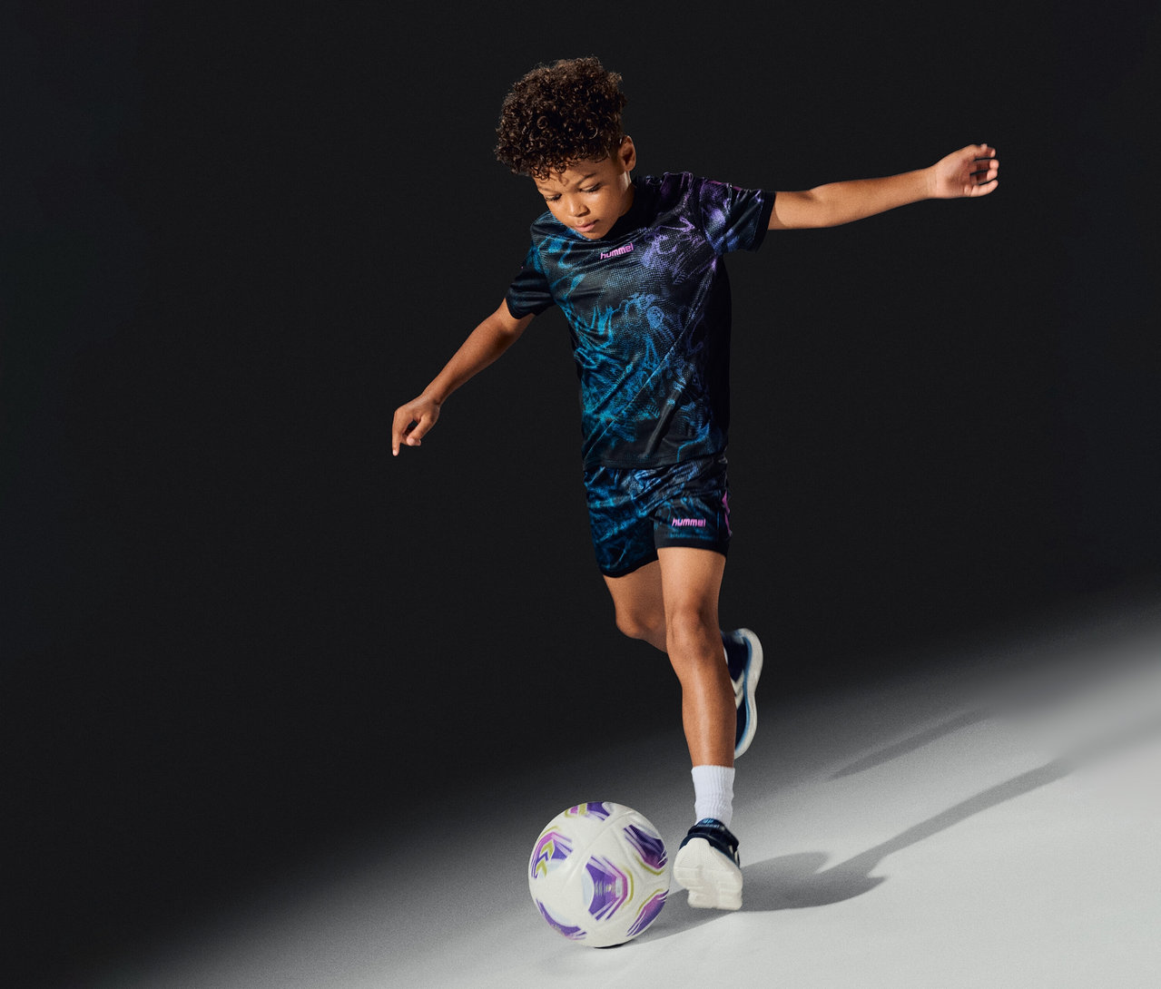 Un garçon portant l'ensemble hummel® « HML Blaze Graphic Set Kids » joue au football, frappant un ballon blanc et violet.