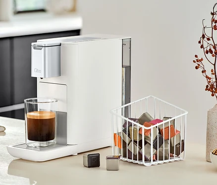 Une machine à café Qbo ESSENTIAL Bright Sand est placée à côté d'un verre de café sur un comptoir de cuisine.