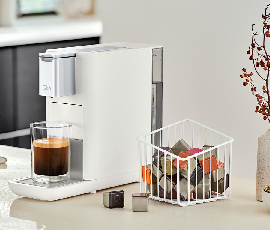 Une machine à café Qbo ESSENTIAL Bright Sand est placée à côté d'un verre de café sur un comptoir de cuisine.