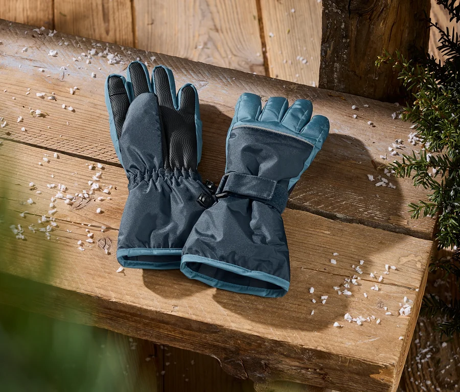 Gants d'hiver bleu foncé avec des accents bleu clair, des poignets élastiques et des sangles ajustables, posés sur une surface en bois avec des flocons de neige et du feuillage vert.