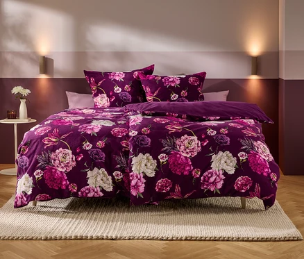 Un lit avec une parure de lit violette à motifs de fleurs roses et blanches se trouve dans une chambre aux murs bicolores et au parquet. Sur une table d'appoint, un vase de fleurs blanches et une bougie.