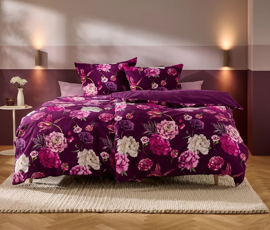 Un lit avec une parure de lit violette à motifs de fleurs roses et blanches se trouve dans une chambre aux murs bicolores et au parquet. Sur une table d'appoint, un vase de fleurs blanches et une bougie.