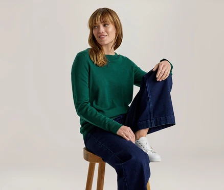 Femme en haut vert à manches longues, jean large bleu foncé et baskets blanches, assise sur un tabouret en bois, regardant à droite et souriant.