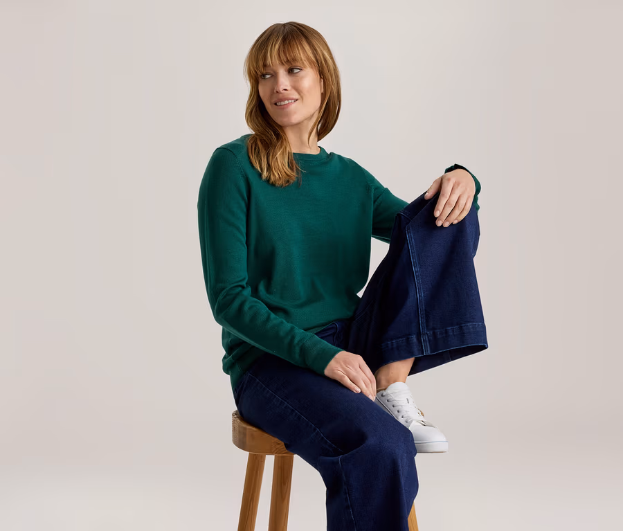 Femme en haut vert à manches longues, jean large bleu foncé et baskets blanches, assise sur un tabouret en bois, regardant à droite et souriant.