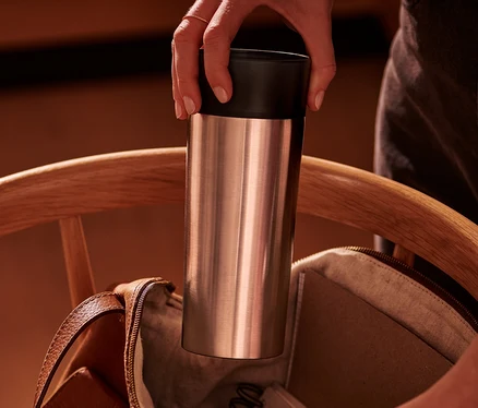 Une main place un gobelet à café to-go en inox dans un sac en cuir marron ouvert sur une chaise en bois.