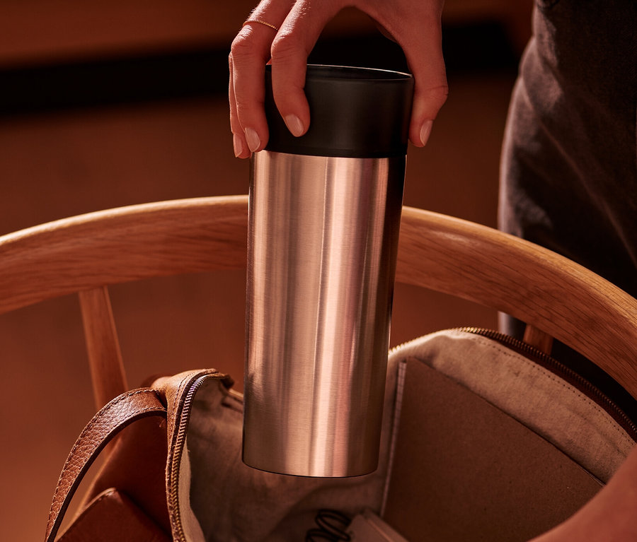 Une main place un gobelet à café to-go en inox dans un sac en cuir marron ouvert sur une chaise en bois.