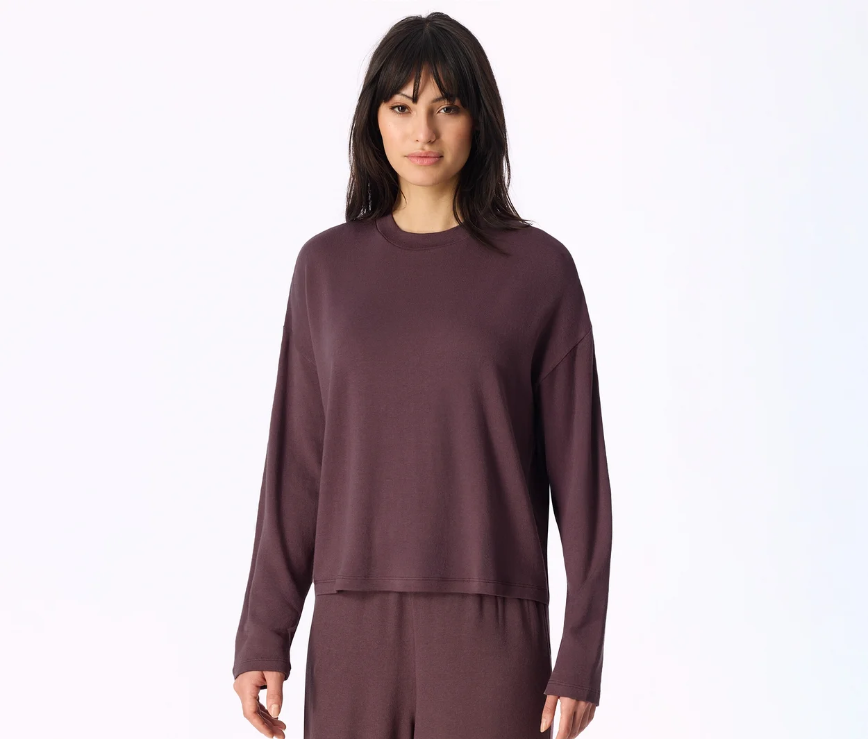 Femme aux cheveux foncés et frange, portant un haut à manches longues et un pantalon assorti de couleur violet foncé, posant devant un fond blanc.