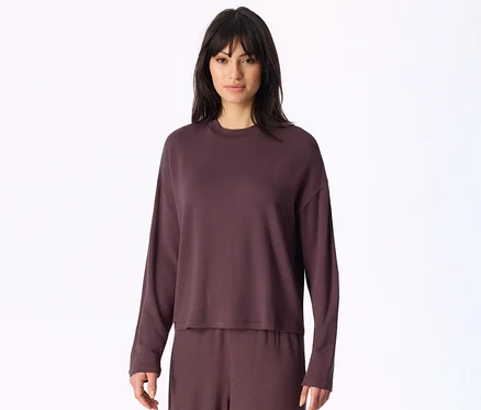 Femme aux cheveux foncés et frange, portant un haut à manches longues et un pantalon assorti de couleur violet foncé, posant devant un fond blanc.