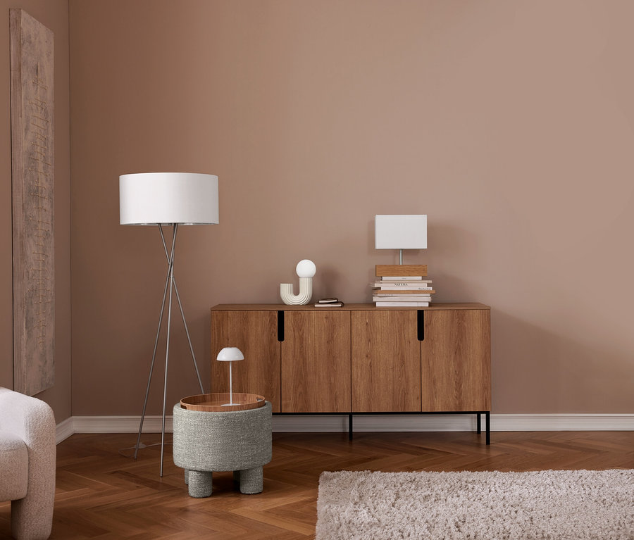Un salon moderne avec des murs bruns et un parquet en chevrons. Un fauteuil de couleur claire est placé à gauche. Un lampadaire blanc à trépied se tient à côté d'une table d'appoint en tissu gris avec un plateau en bois et une petite lampe blanche. Au centre, une commode en bois à quatre portes supporte des piles de livres, une lampe globe blanche, une décoration blanche incurvée et une lampe de table blanche rectangulaire. Un tapis shaggy de couleur claire est posé sur le sol devant la commode.