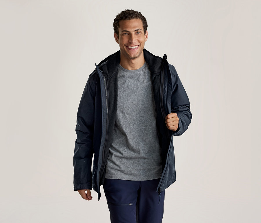Un homme souriant porte une veste bleu foncé sur un t-shirt gris et un pantalon bleu foncé. Il tient la veste ouverte avec sa main droite.