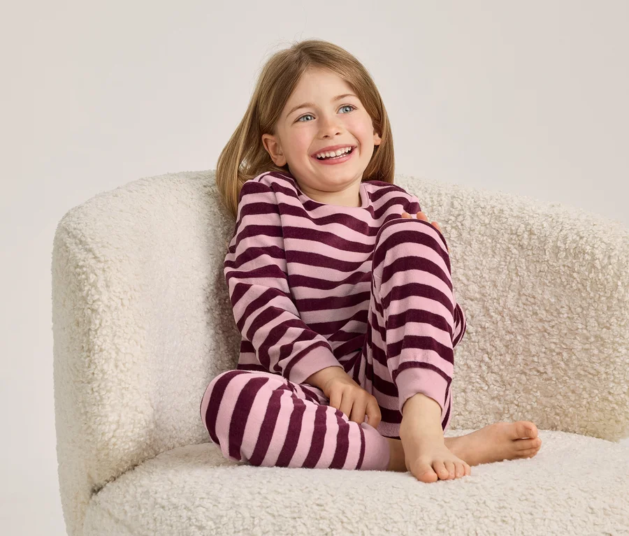 Jeune fille aux cheveux blonds et aux yeux bleus, souriante, assise pieds nus dans un pyjama rayé sur un fauteuil crème.