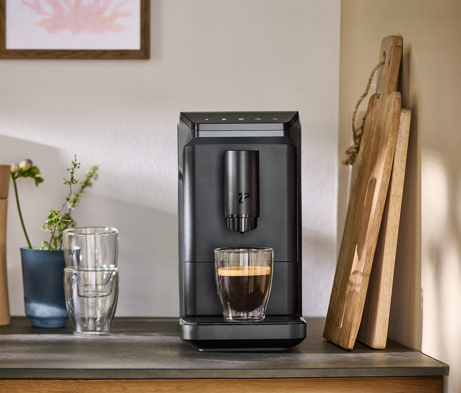 Machine à café entièrement automatique « Esperto mini » grise verse du café dans un verre, à côté de planches à découper, moulins à épices et verres sur un comptoir.