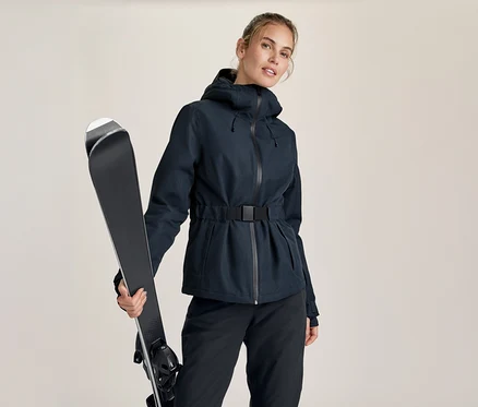 Femme en veste de ski bleu foncé ceinturée et pantalon sombre, tenant une paire de skis noirs sur son épaule gauche.