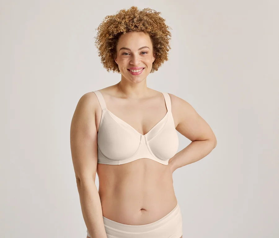 Une femme souriante aux cheveux bouclés, vêtue d'un soutien-gorge de couleur claire et d'un slip assorti, se tient la main sur la hanche.