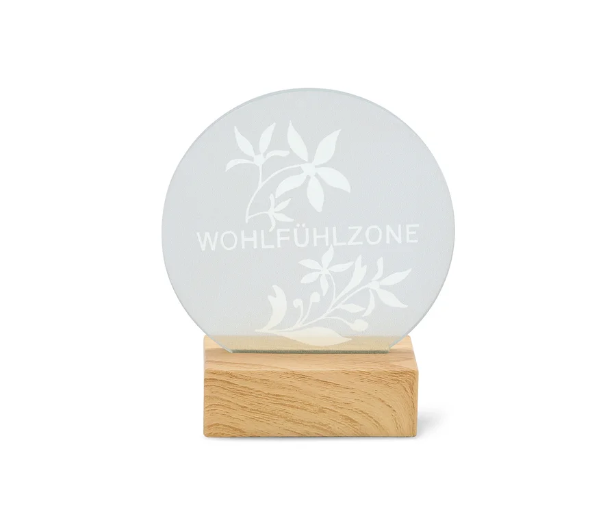 Panneau rond translucide avec l'inscription "WOHLFÜHLZONE" et des motifs floraux, posé sur une base en bois clair.