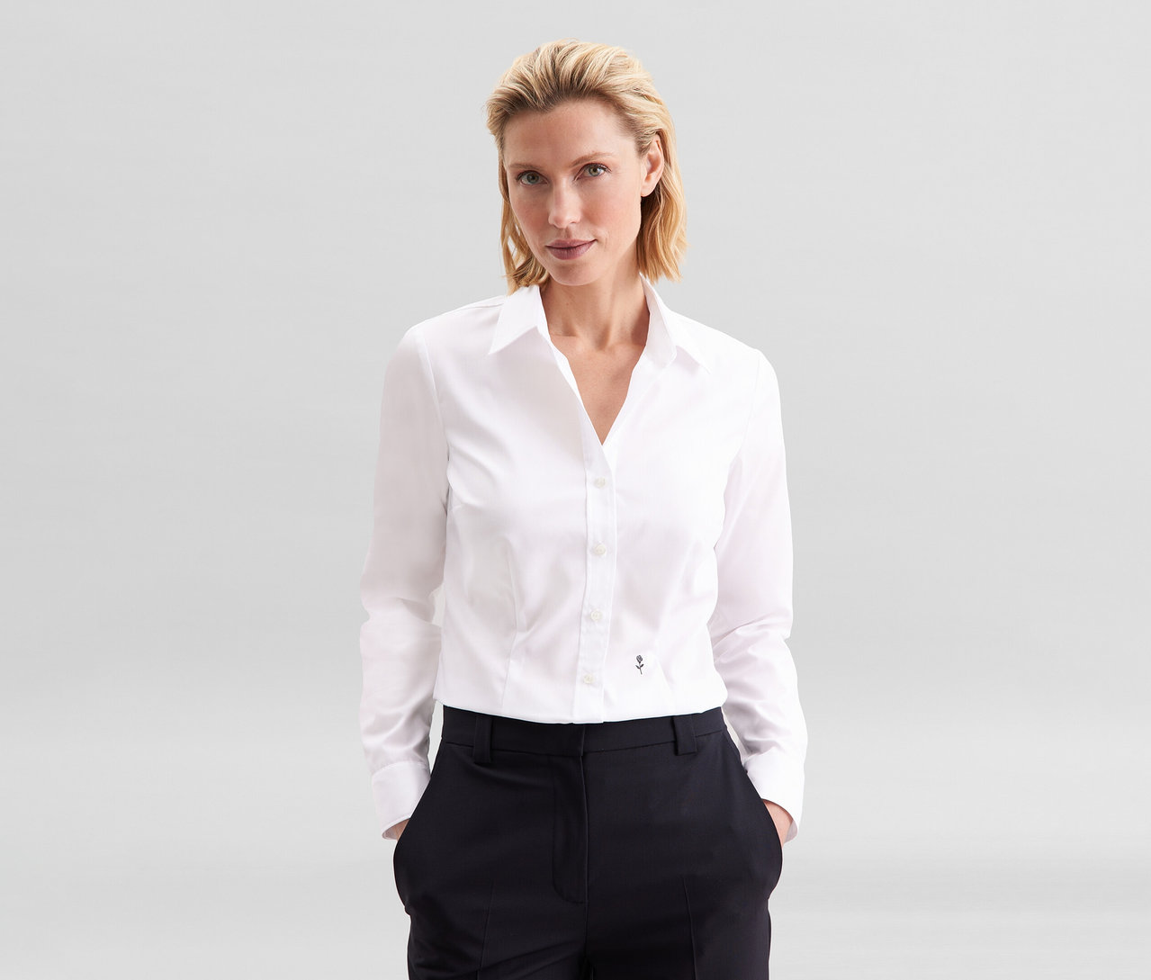 Femme blonde portant un chemisier Slim Fit non iron Seidensticker blanc et un pantalon foncé, les mains dans les poches.