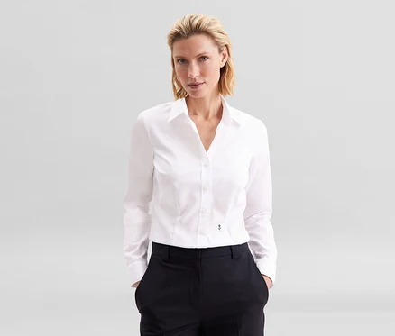 Femme blonde portant un chemisier Slim Fit non iron Seidensticker blanc et un pantalon foncé, les mains dans les poches.