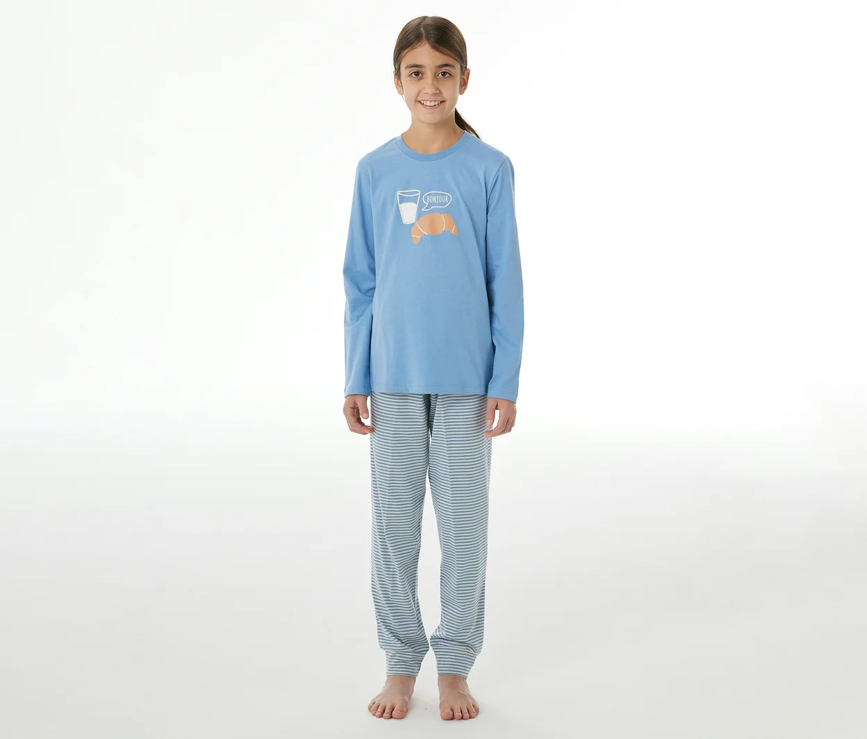 Jeune fille portant un pyjama enfant bleu à manches longues et un pantalon rayé. Le haut présente un verre de lait et un croissant avec l'inscription « Bonjour ».