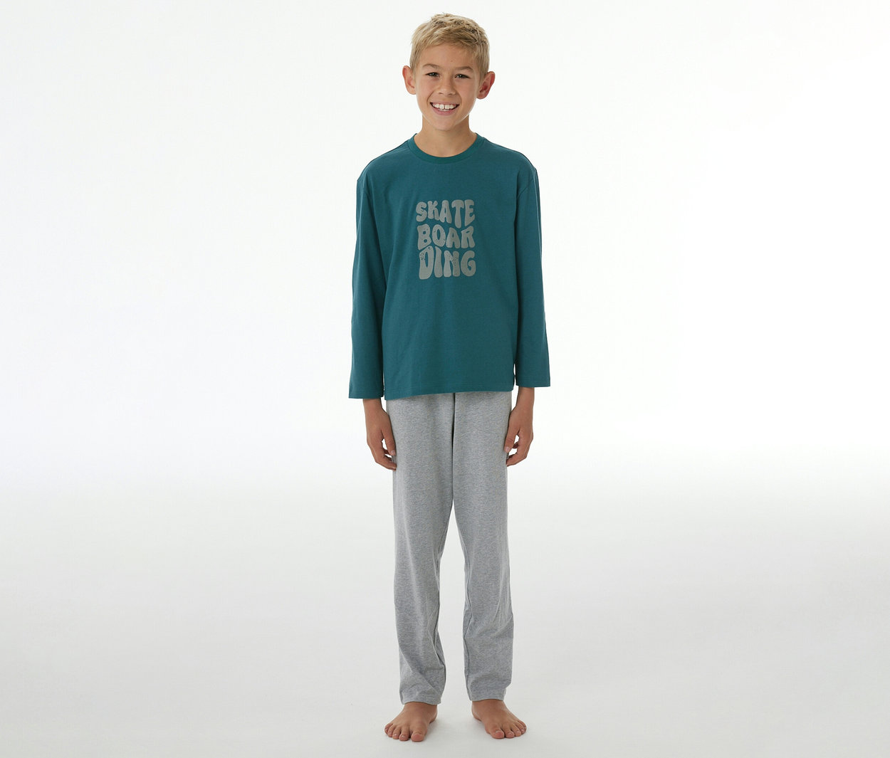Un garçon souriant se tient pieds nus, portant un pyjama SCHIESSER Kinder-Pyjama »Skate« composé d'un t-shirt à manches longues vert foncé avec l'inscription "SKATE BOARDING" et d'un pantalon gris.