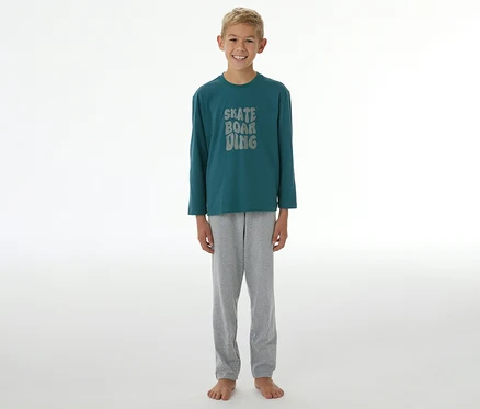 Un garçon souriant se tient pieds nus, portant un pyjama SCHIESSER Kinder-Pyjama »Skate« composé d'un t-shirt à manches longues vert foncé avec l'inscription "SKATE BOARDING" et d'un pantalon gris.